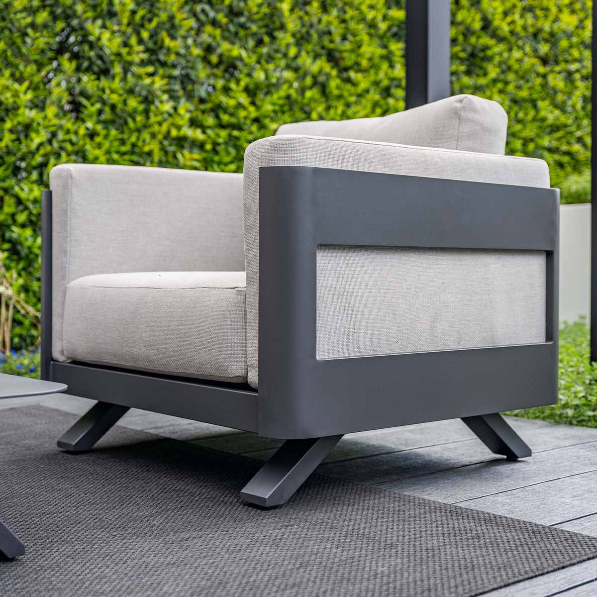 Grijze fauteuil met donker metalen frame op houten dek, achter groene heg.