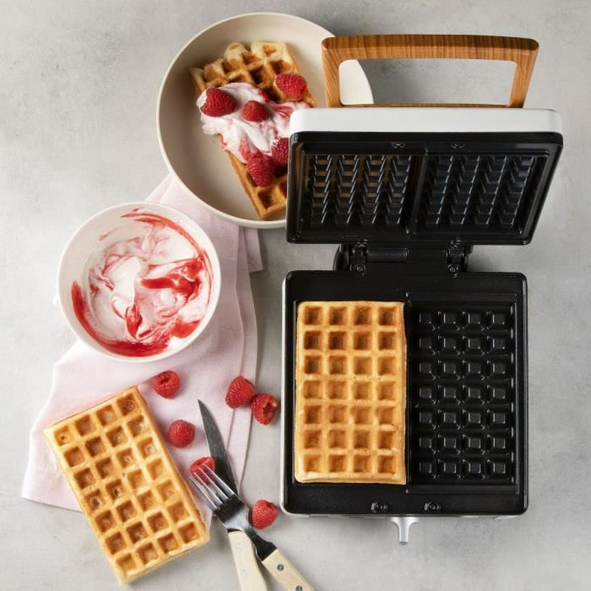 Ein Waffeleisen mit einem fertigen Waffel und einer leeren Form, daneben Waffeln mit Beeren und Sahne.