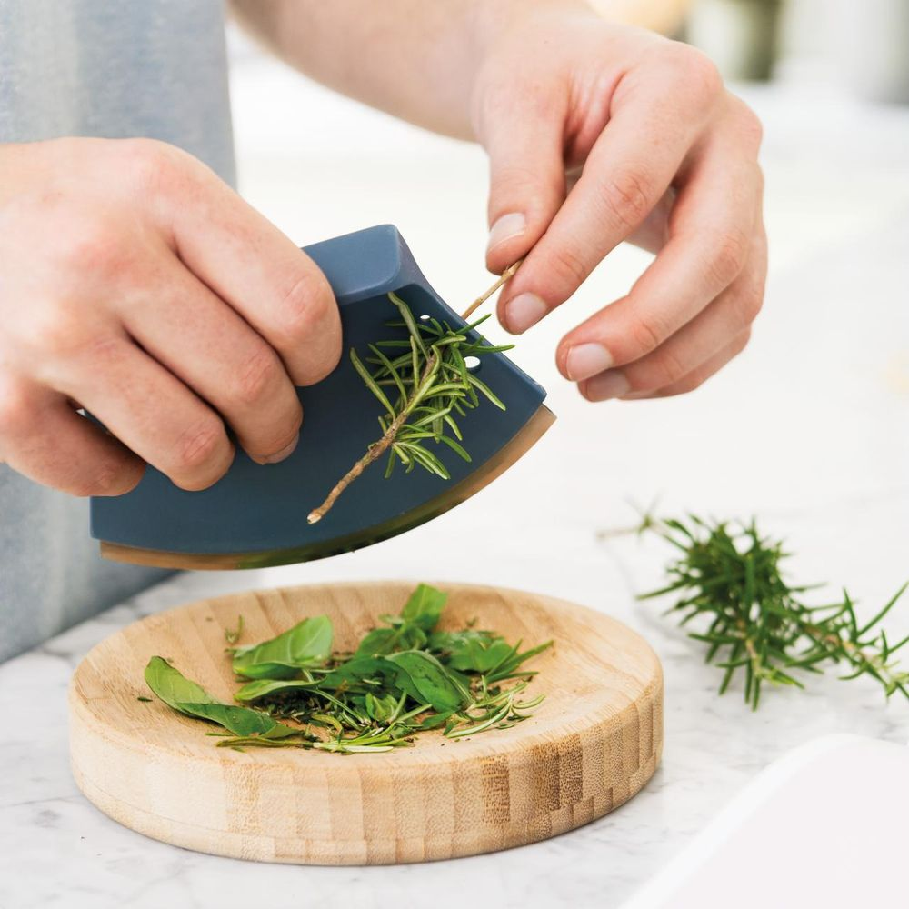 Persoon hakt kruiden met een blauw mezzaluna mes in een houten kom.