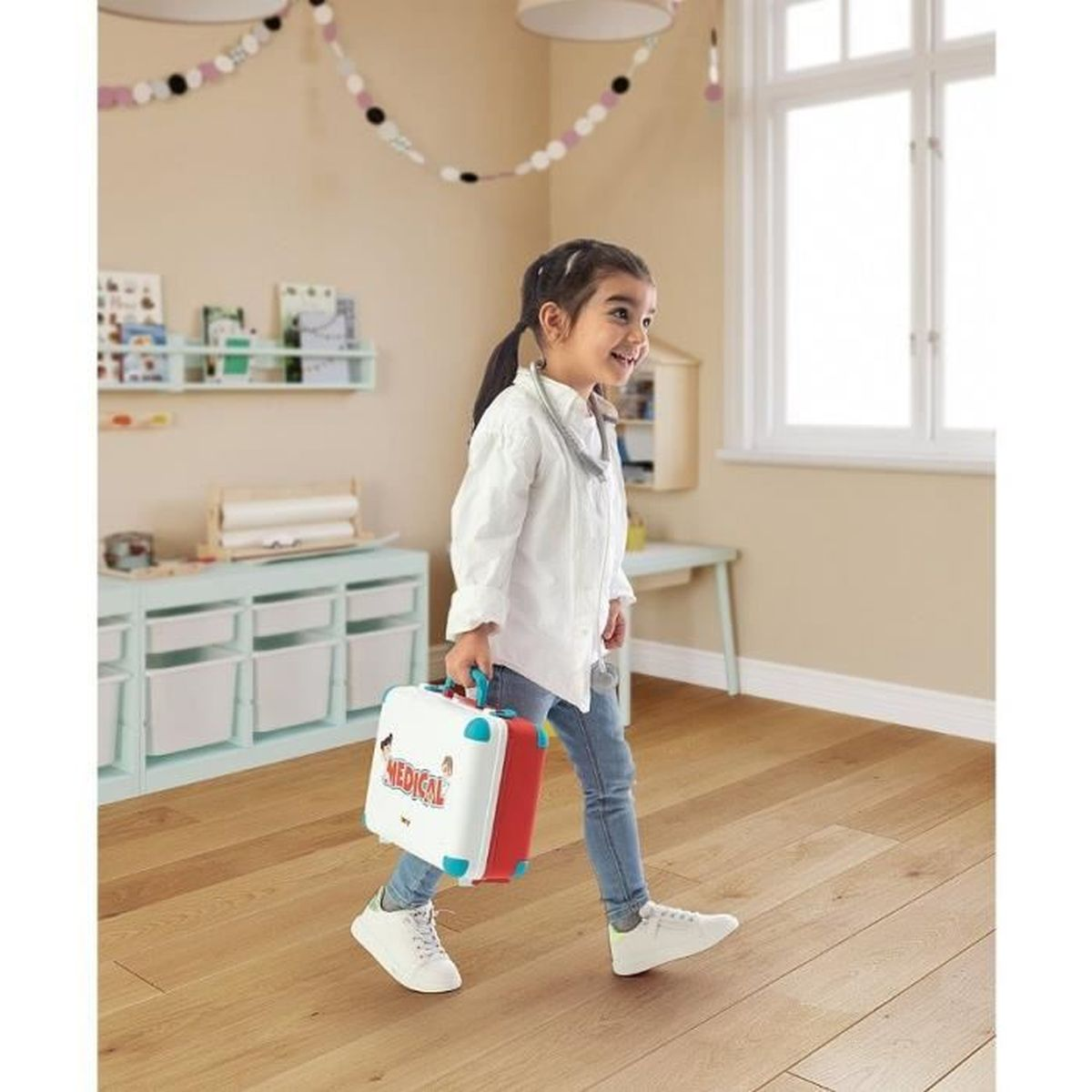 Una niña con bata de doctora lleva un maletín médico blanco y rojo.