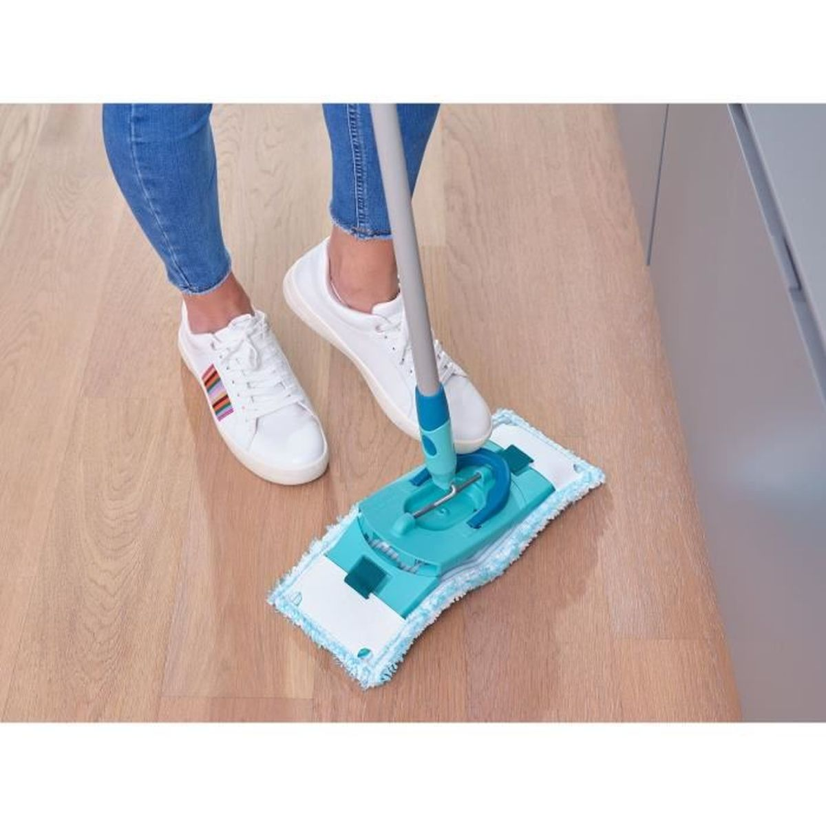 Una persona lava un pavimento in legno con uno spazzolone blu. Scarpe da ginnastica bianche visibili.