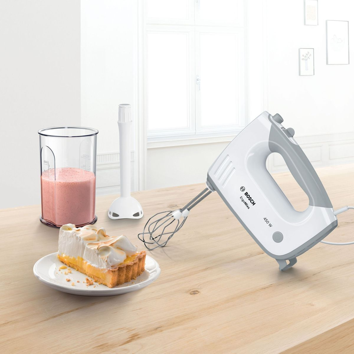 A white Bosch hand mixer with a gray handle on a wooden table next to a glass of smoothie and cake.