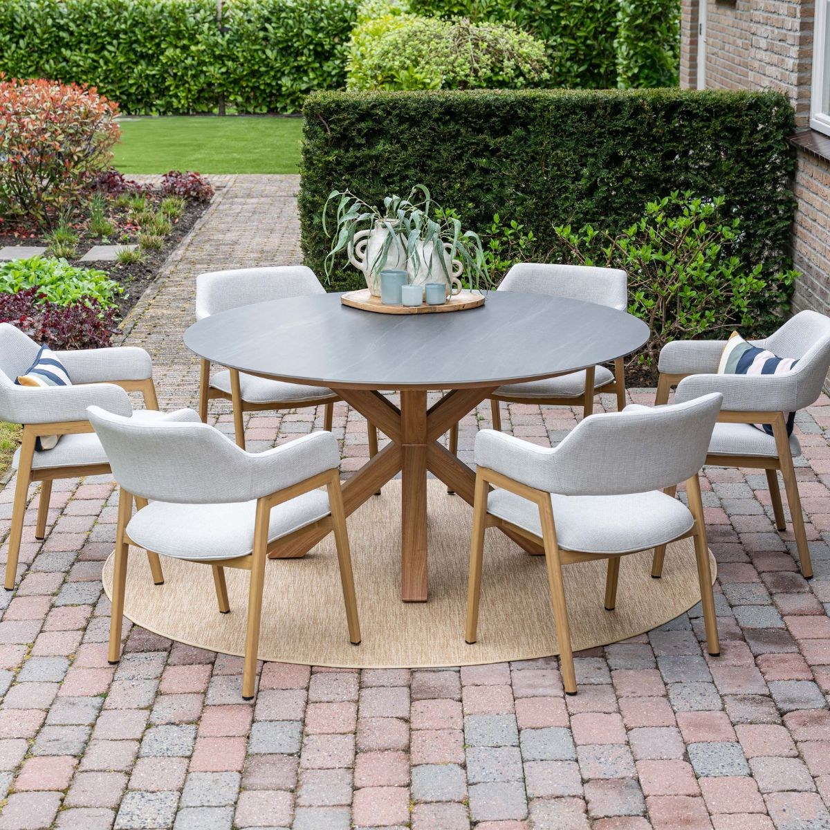 Een ronde tafel met zes stoelen op een betegeld terras, omringd door groen.