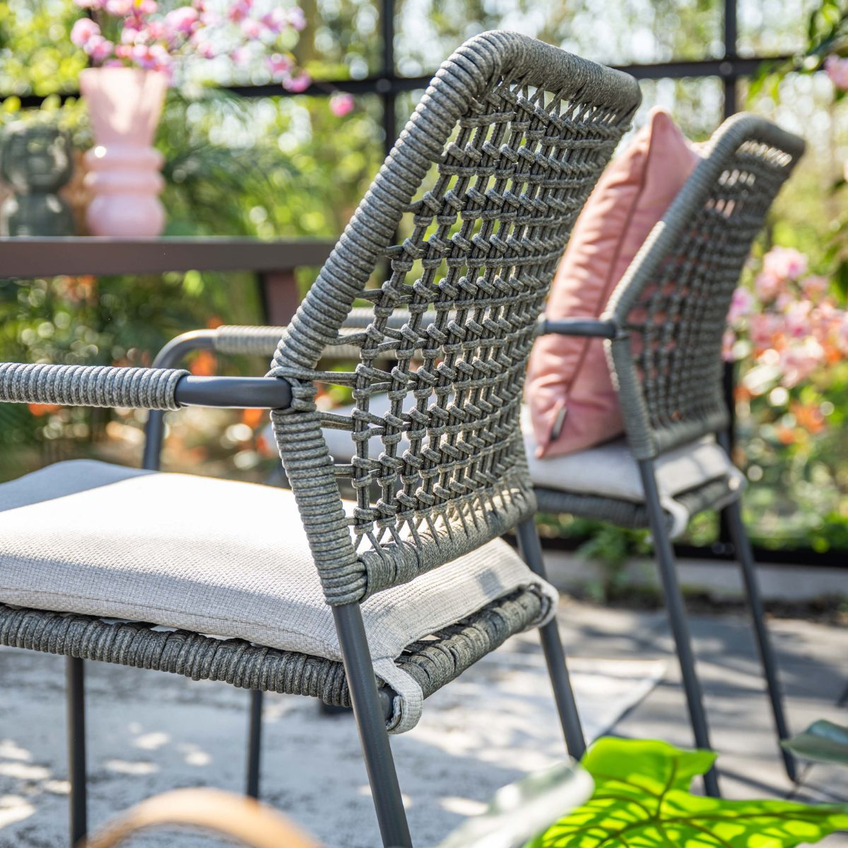 Twee grijze rotan stoelen met kussens en roze kussens in een tuin.