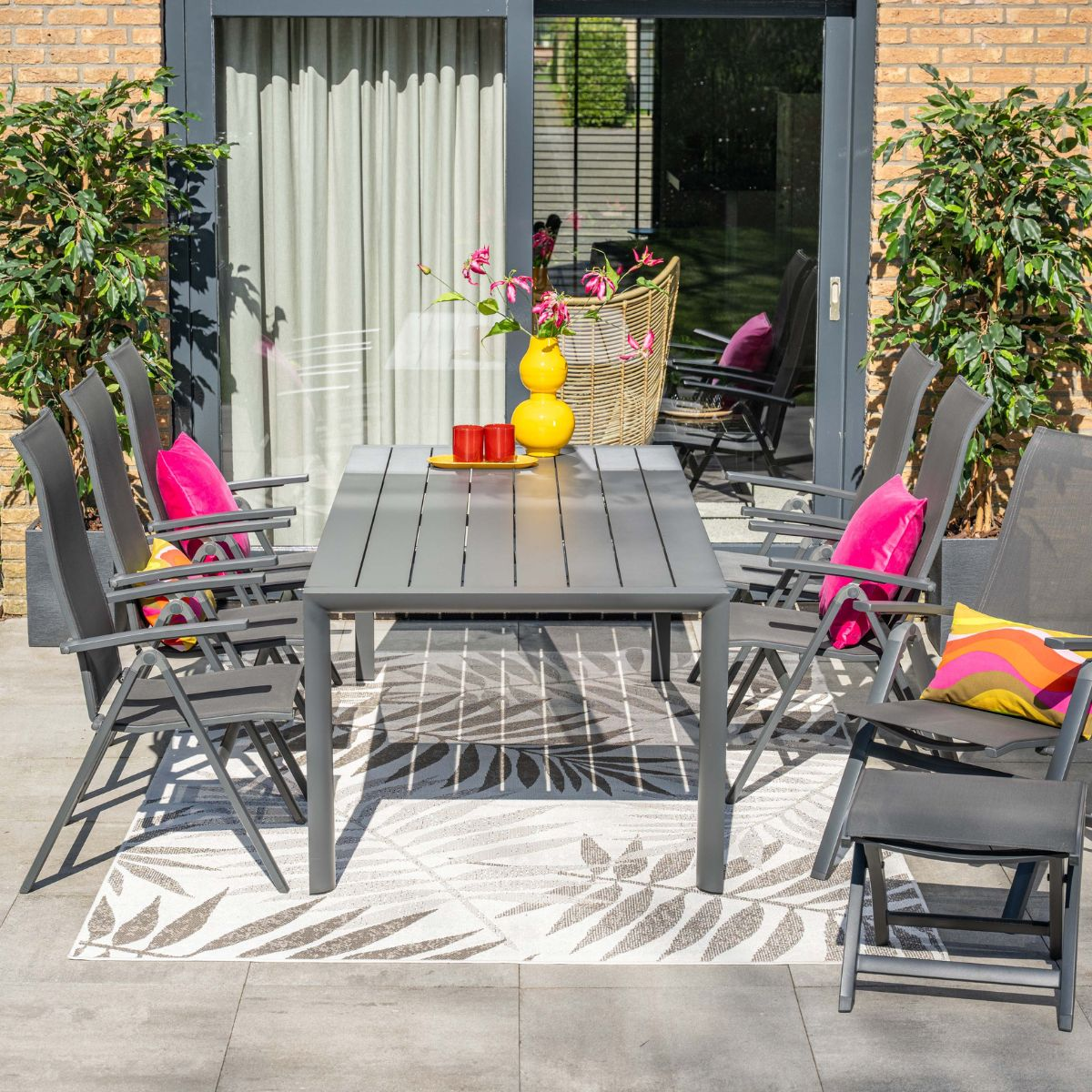 Grijze tuintafel met stoelen, kleurrijke kussens en gele vaas.