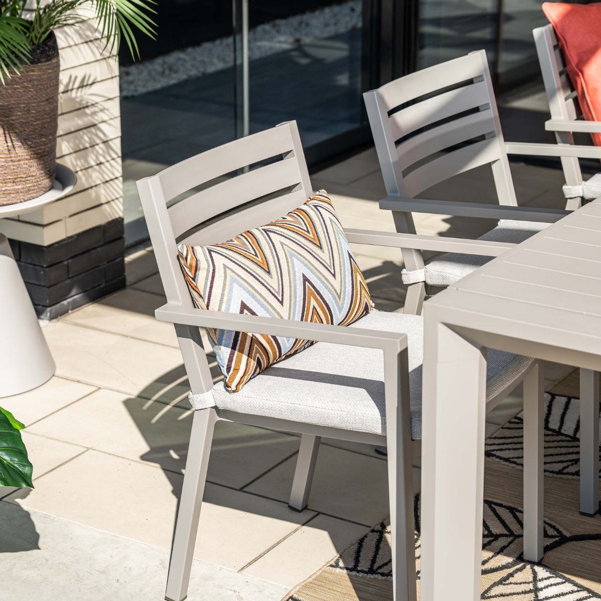 Grijze buitenstoelen met een patroonkussen op een betegeld terras.