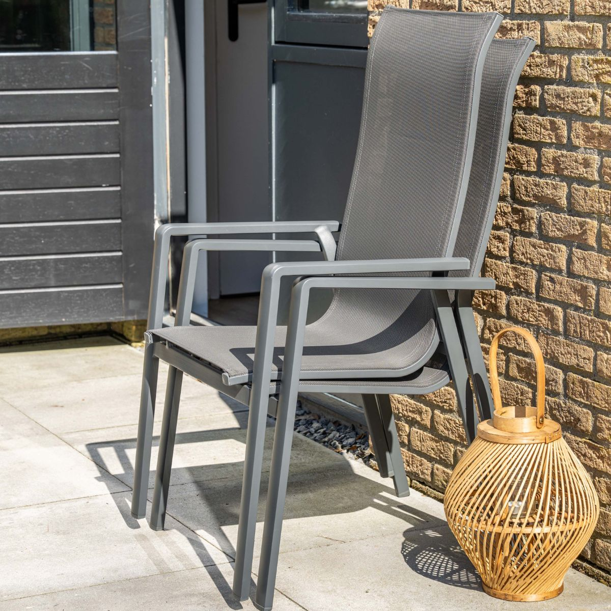 Twee grijze tuinstoelen op een terras naast een bakstenen muur en een lantaarn.