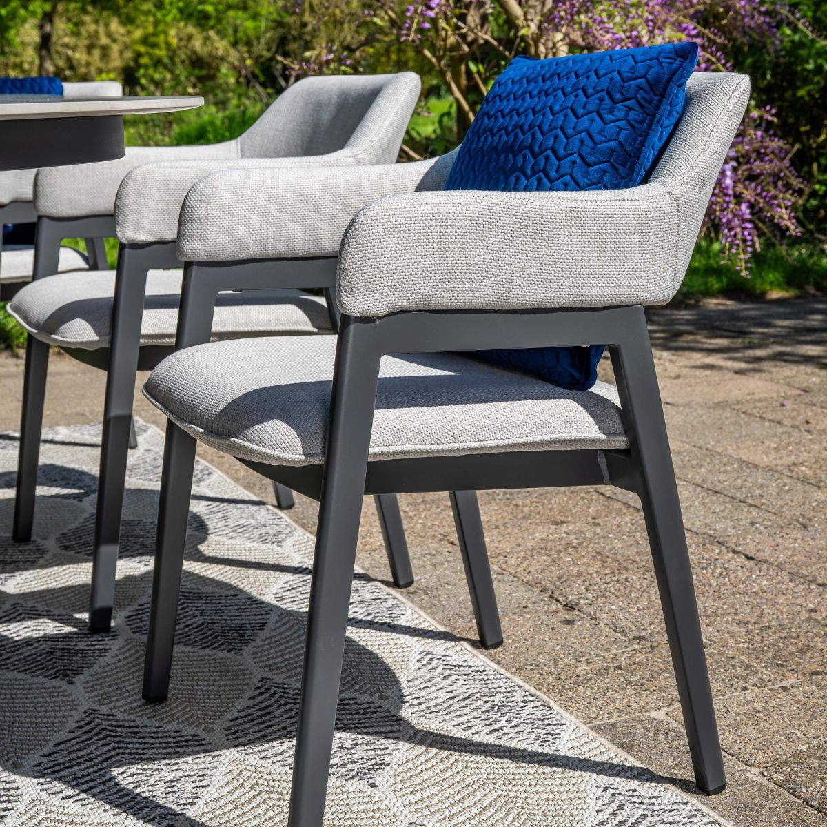 Grijze tuinstoelen met blauwe kussens op een kleed, met paarse bloemen achtergrond.
