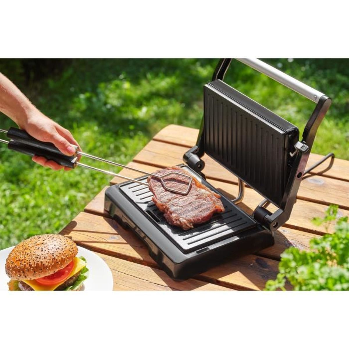 Carne su una griglia, accanto a un hamburger su un tavolo all'aperto.