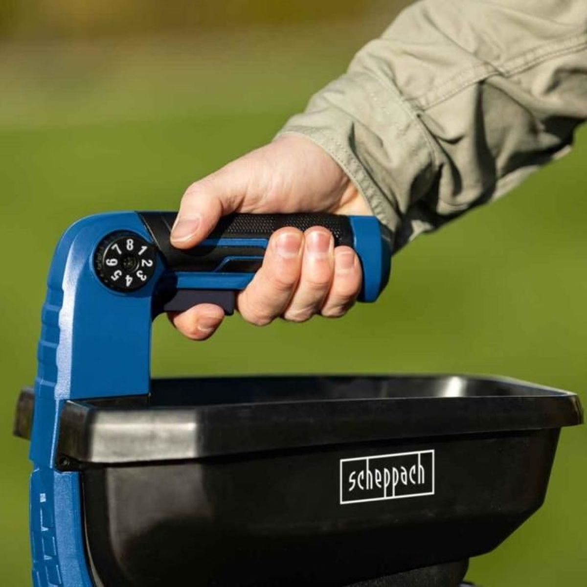 Una mano tiene uno spargitore di fertilizzante blu e nero con un quadrante numerato da 1 a 8.