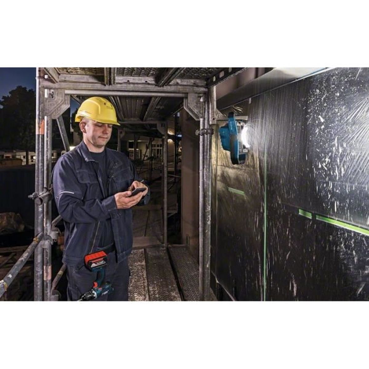 Bauarbeiter in gelbem Helm prüft Smartphone auf Gerüst. Blaue Lampe leuchtet.