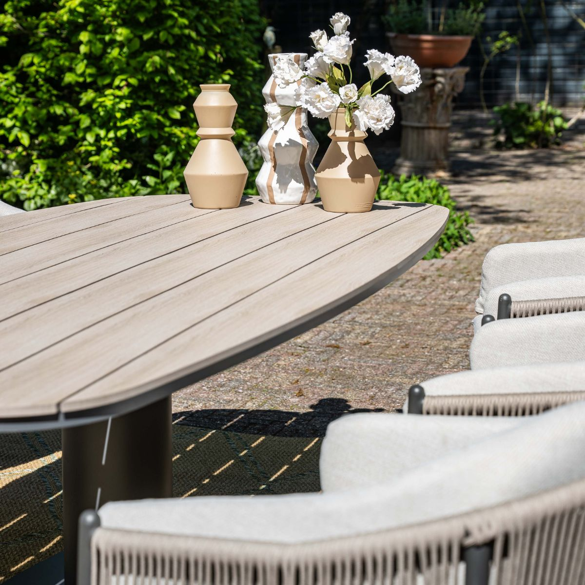 Houten tafel met vazen en bloemen buiten, omringd door stoelen en groen.
