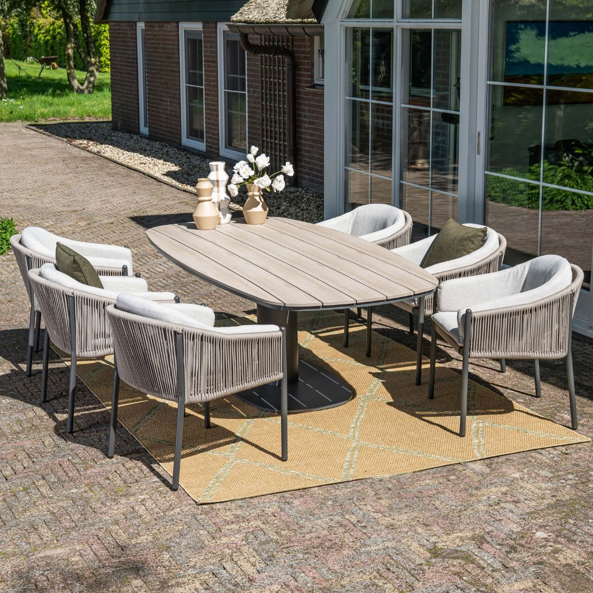 Een ovale houten tafel met zes stoelen op een patio met een kleed.