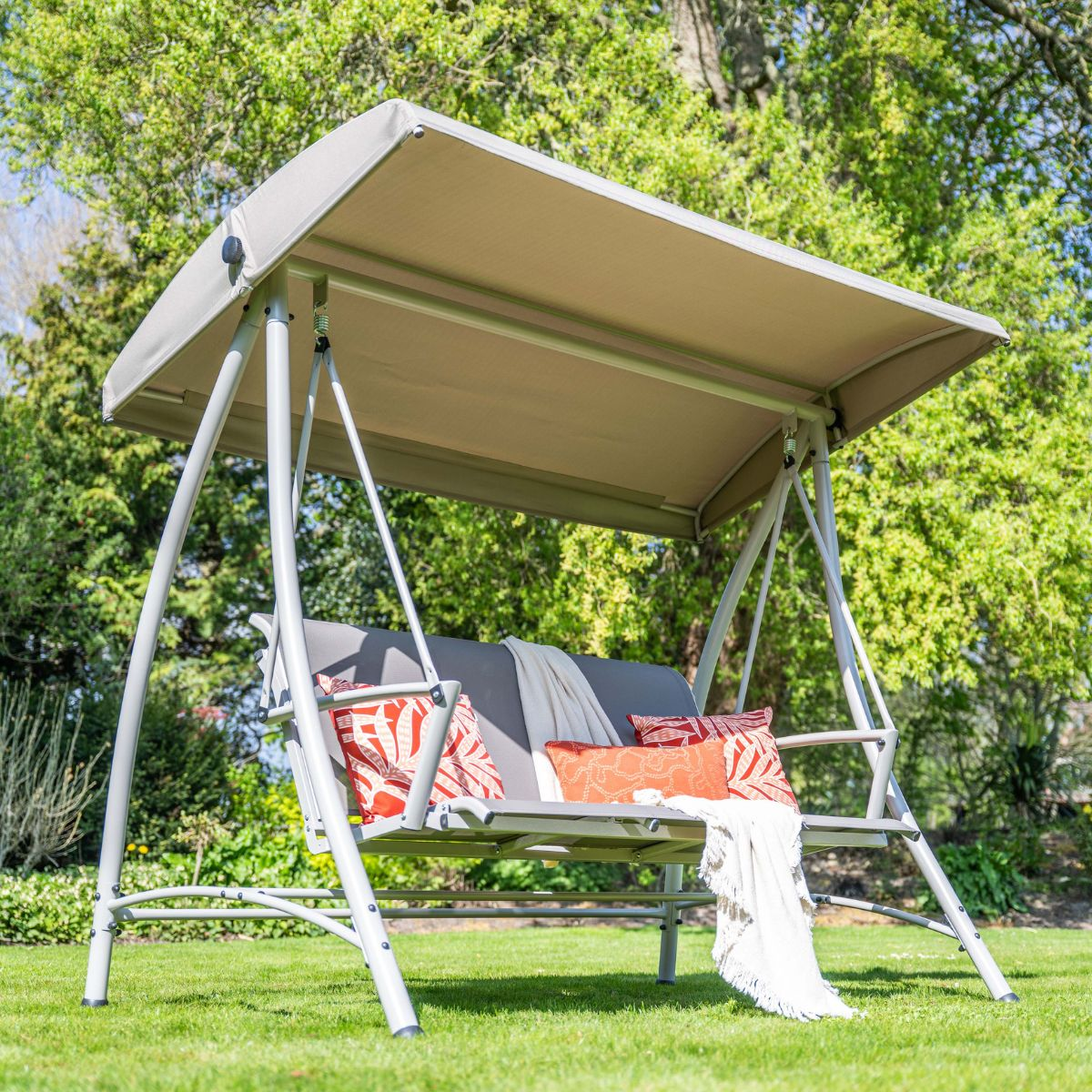 Een beige canopy schommel met grijze kussens en decoratieve kussens op een groen gazon.