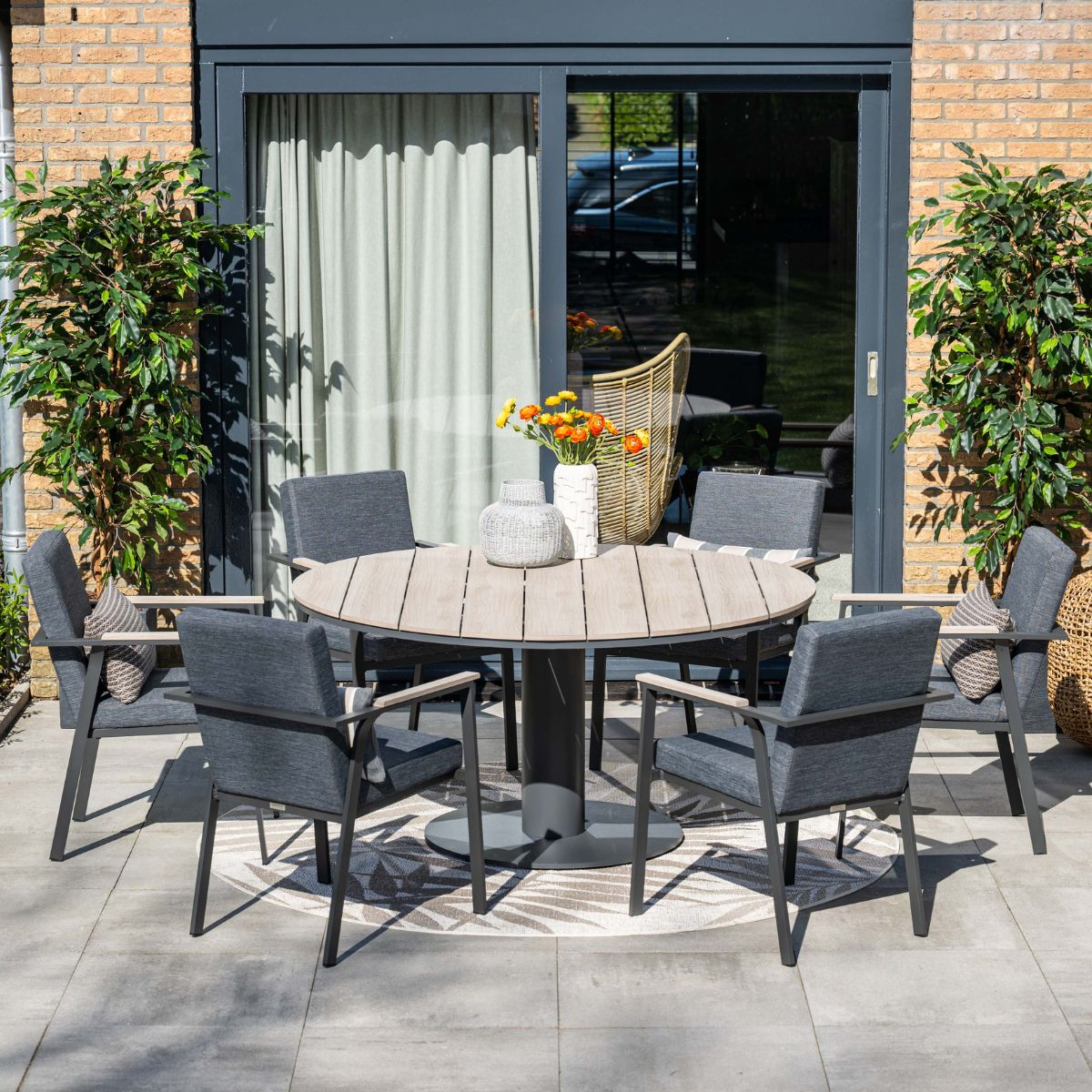 Ronde buitentafel met zes stoelen, omringd door planten en een glazen deur.