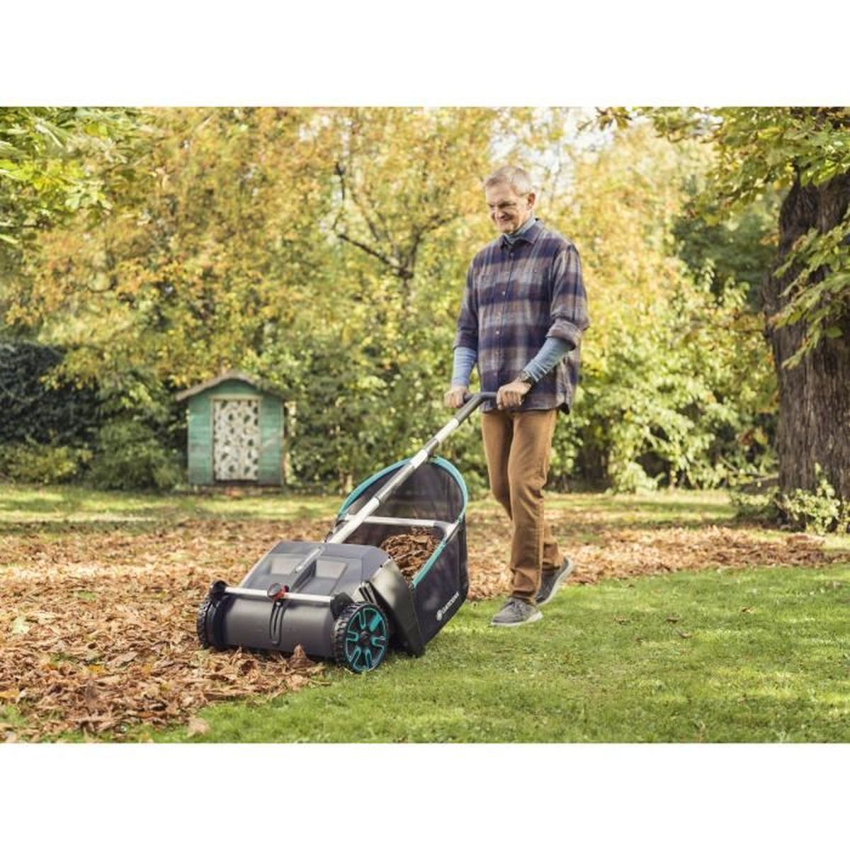 Mann schiebt einen Laubsammler durch Herbstlaub vor einem kleinen Schuppen.