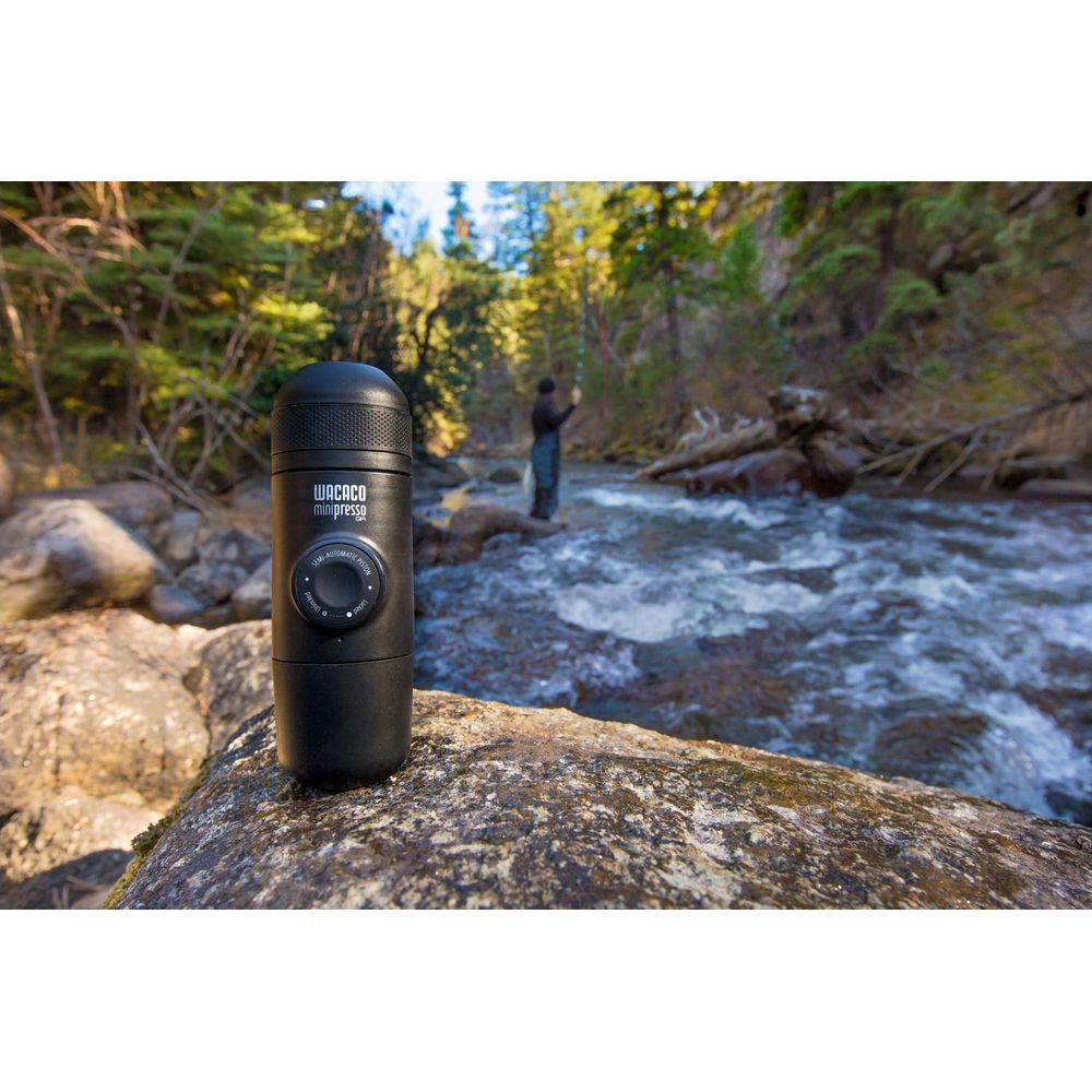 Ein schwarzes Wacaco Minipresso liegt auf einem Felsen neben einem Fluss mit einem Angler im Hintergrund.