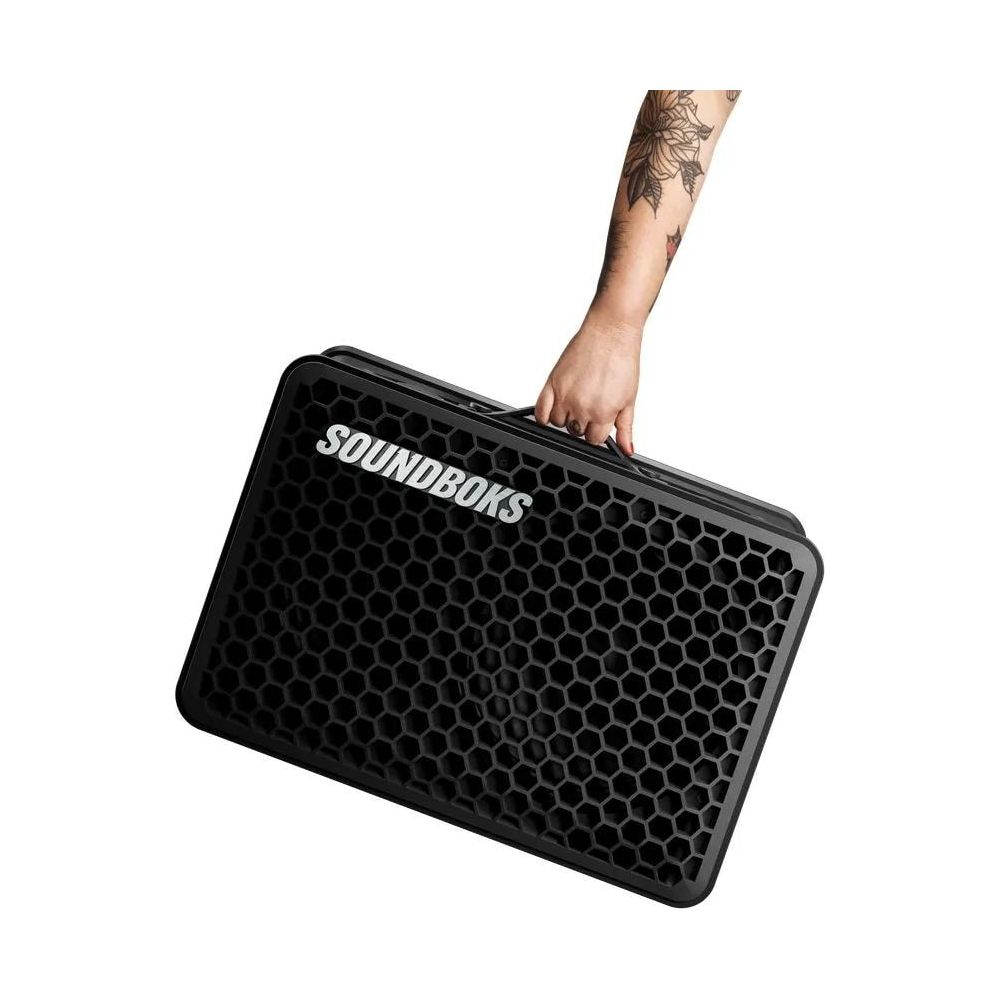 Eine Hand mit Tätowierung trägt einen schwarzen Lautsprecher mit der Aufschrift SOUNDBOKS.