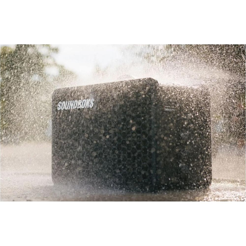 Ein dunkler Lautsprecher mit dem Logo SOUNDBOKS wird von Wassersprühstrahlen getroffen.