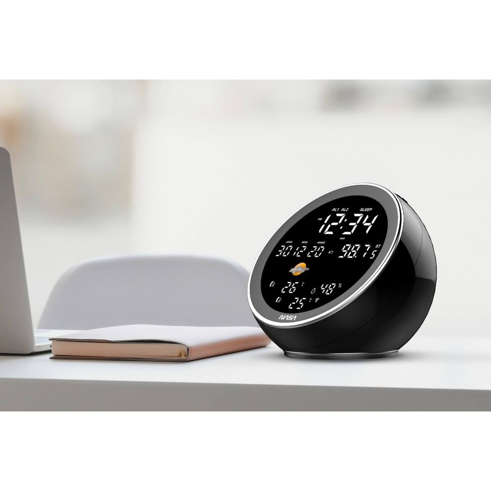 Czarny zegar cyfrowy pokazujący datę, temperaturę i wilgotność na biurku.