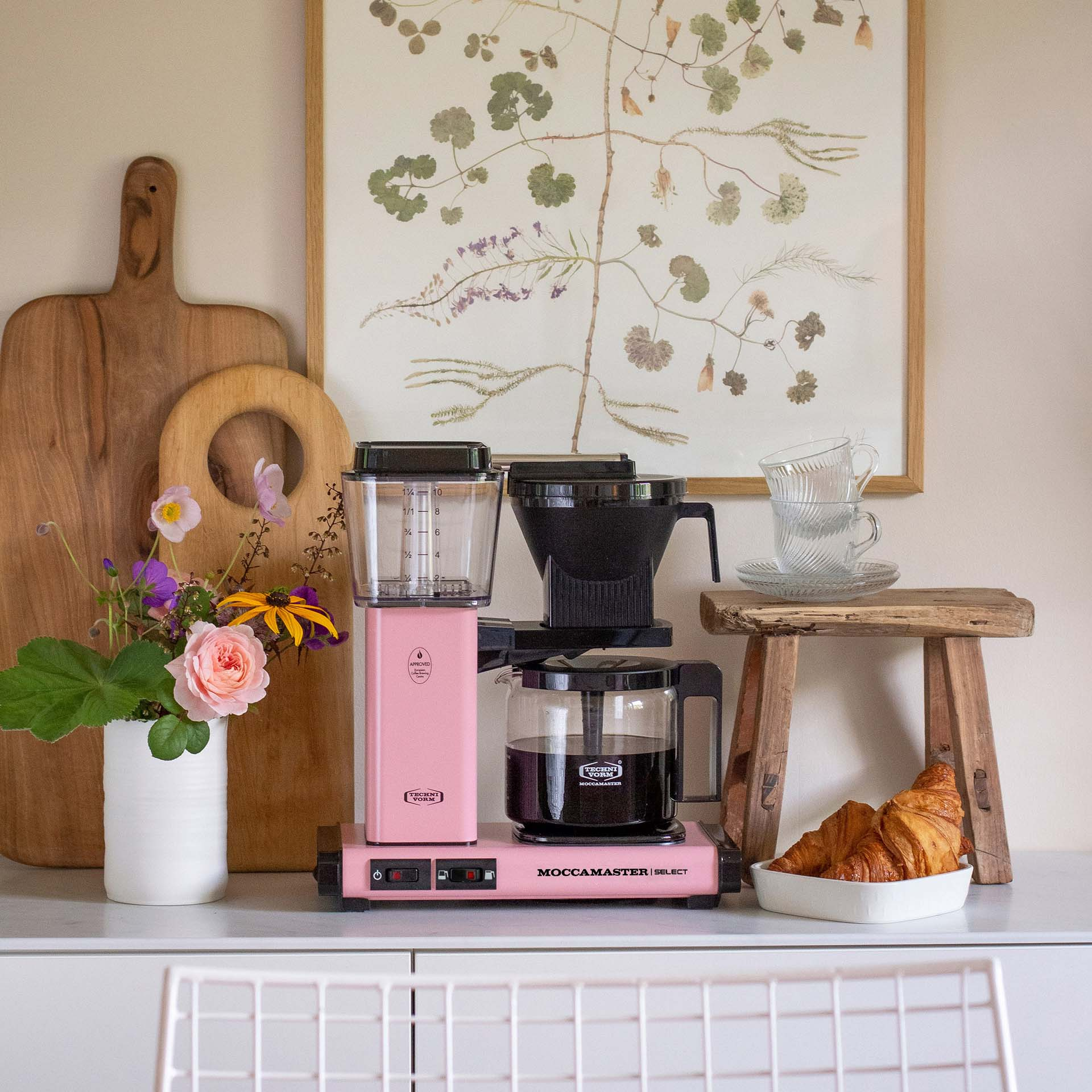 Eine rosa Kaffeemaschine mit Kanne, Blumen, Holzbrettern und Croissants.