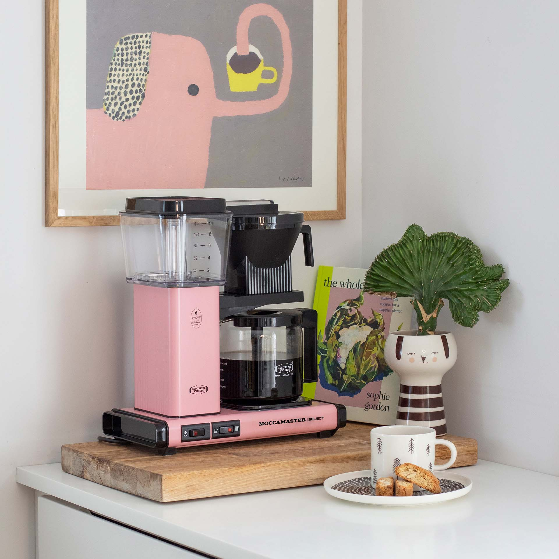 Eine rosa Kaffeemaschine steht auf einem Holzbrett. Daneben ein Buch, eine Pflanze und eine Tasse.
