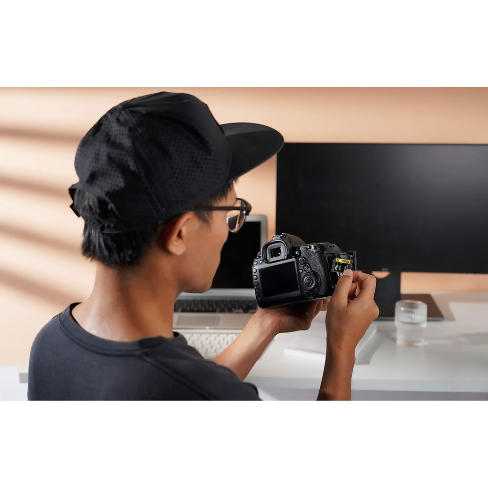 A man wearing a black cap and glasses holds a camera, inserting a memory card.