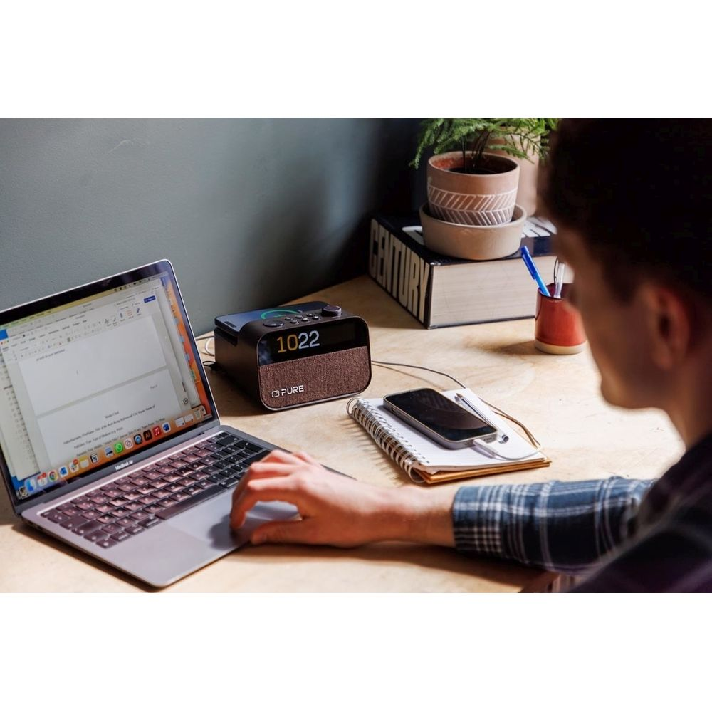 Hombre trabajando en portátil; radio digital, teléfono y cuaderno en escritorio.