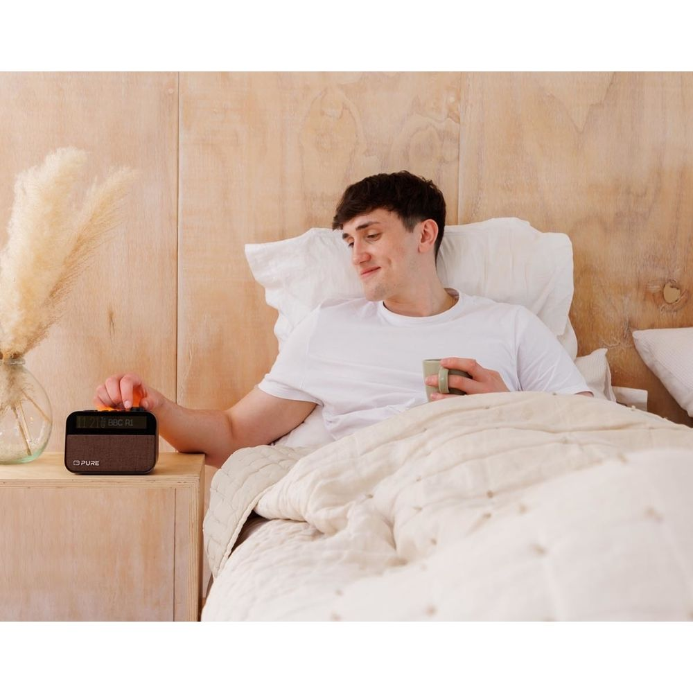Un hombre en la cama se acerca a una radio, sosteniendo una taza.