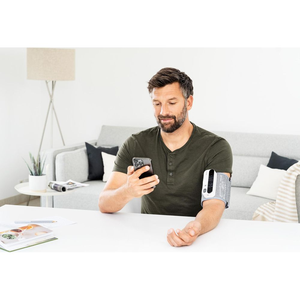 Ein Mann sitzt an einem Tisch und benutzt ein Blutdruckmessgerät am Arm und ein Smartphone.