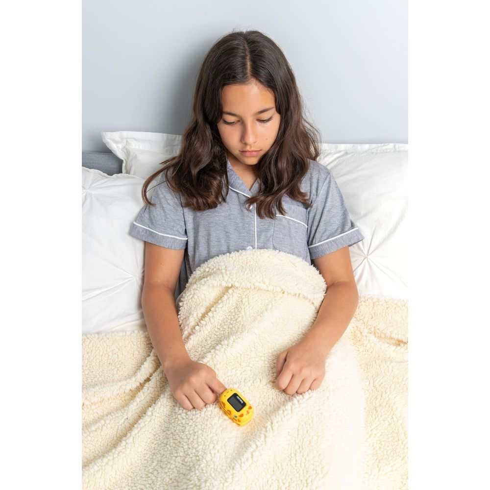 Mädchen im Bett mit Pulsoximeter in der Hand, unter einer cremefarbenen Decke.