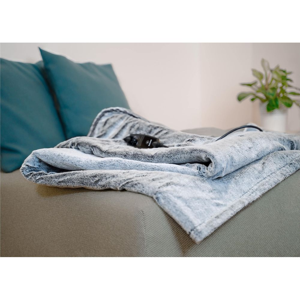 Graue Heizdecke mit schwarzem Regler auf einem Sofa, neben zwei Kissen.