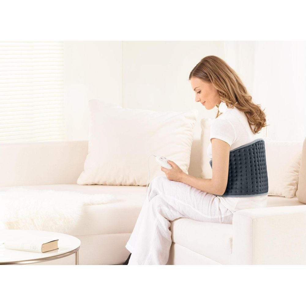 Eine Frau sitzt auf einem Sofa mit einer Heizmatte am Rücken und hält eine Fernbedienung.