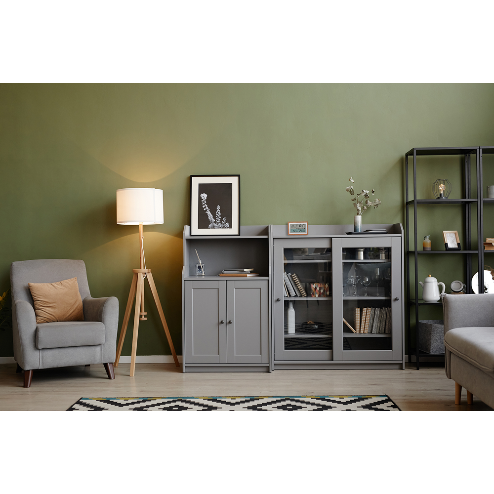 Ein grauer Sessel, ein Stehlampen und ein Schrank mit Glasfront in einem grünen Raum.
