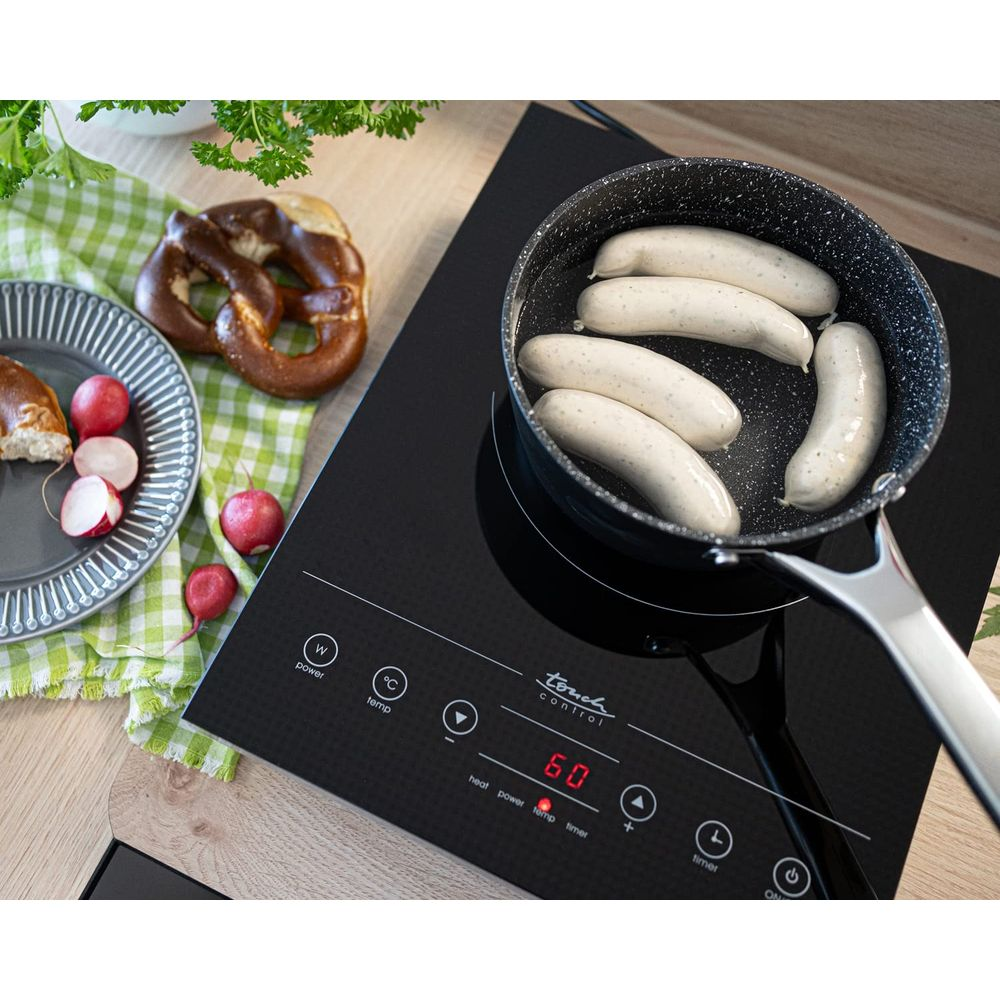 Pięć kiełbasek smaży się na patelni na płycie indukcyjnej. W pobliżu znajdują się precle i rzodkiewki.