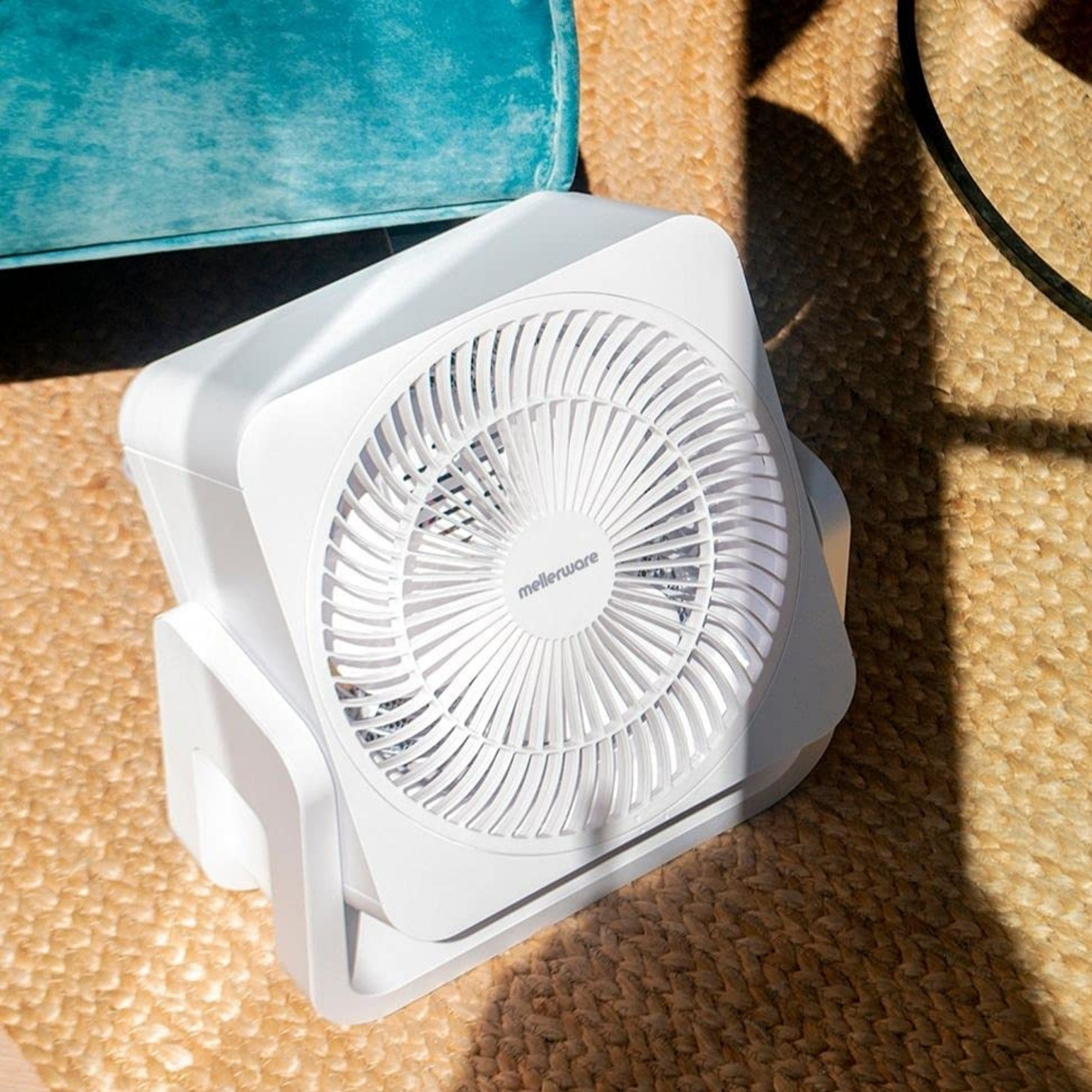 Un ventilador de caja blanco sobre una alfombra de yute junto a un sofá turquesa.