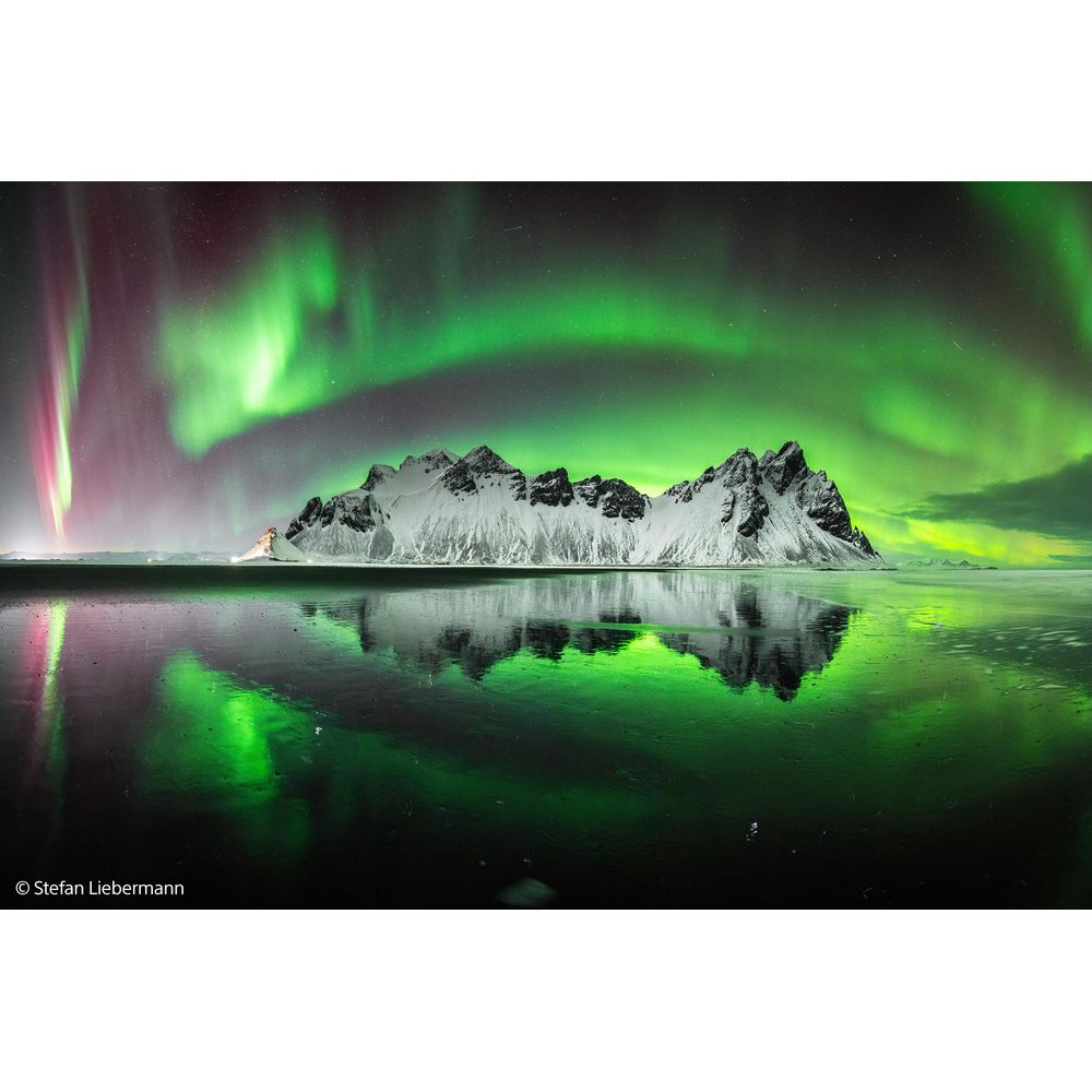 Snowy mountains reflect in the water under the Northern Lights.