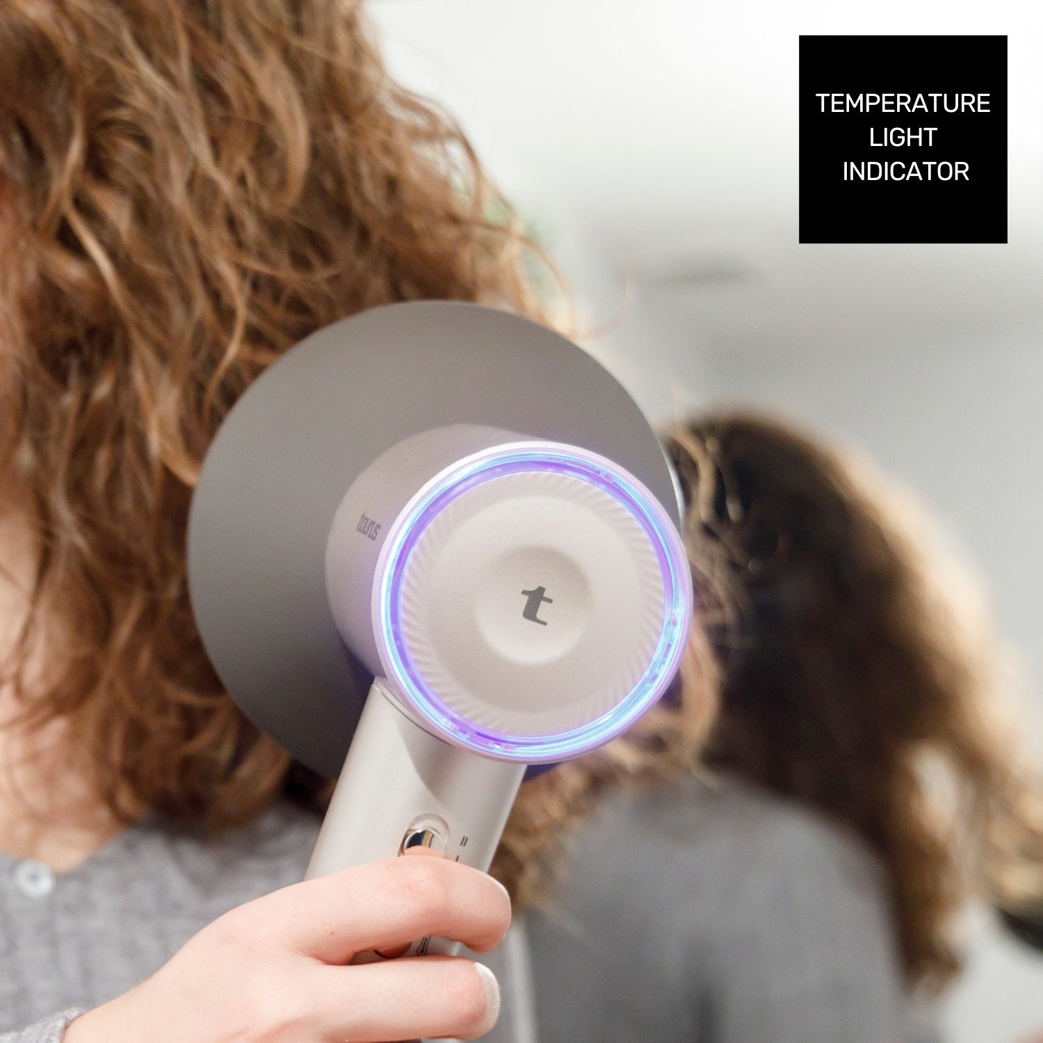 Una mano sostiene un secador de pelo con anillo de luz azul-púrpura frente a cabello castaño.