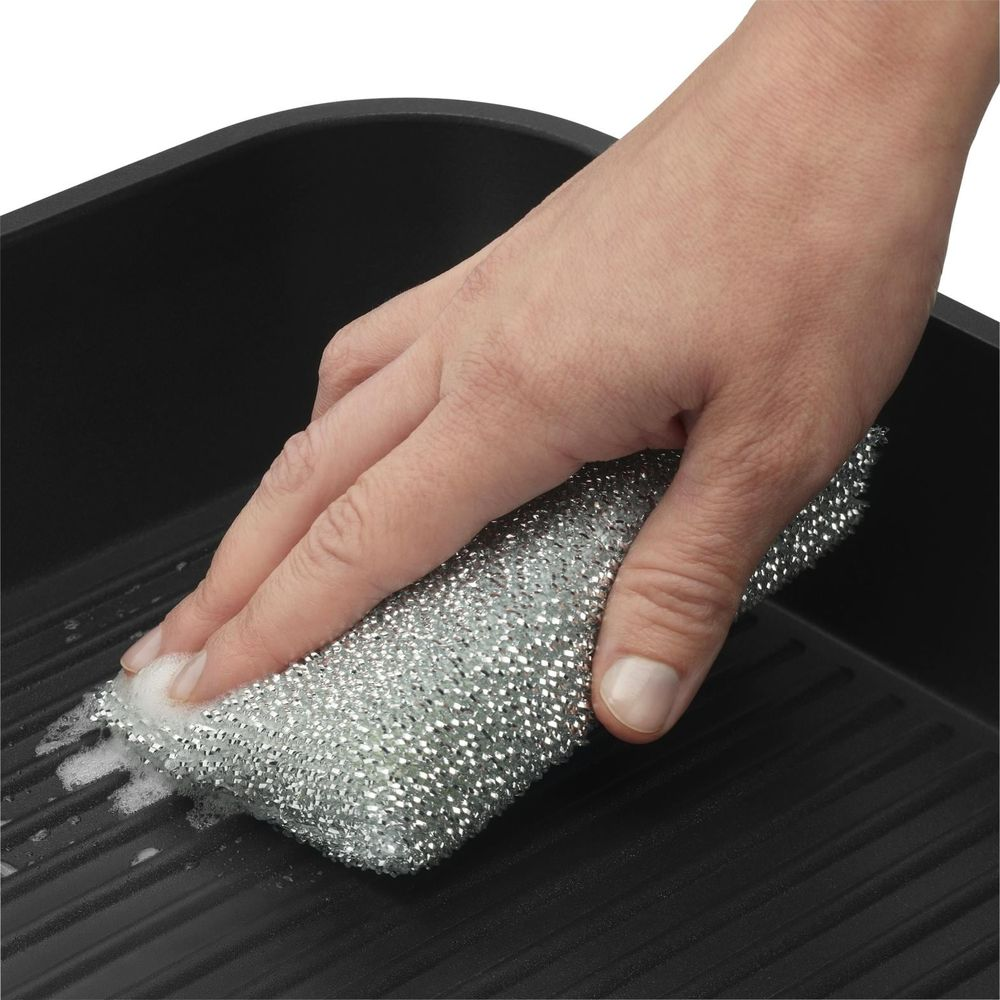 Eine Hand reinigt eine Grillpfanne mit einem silbernen Schwamm und Seifenblasen.