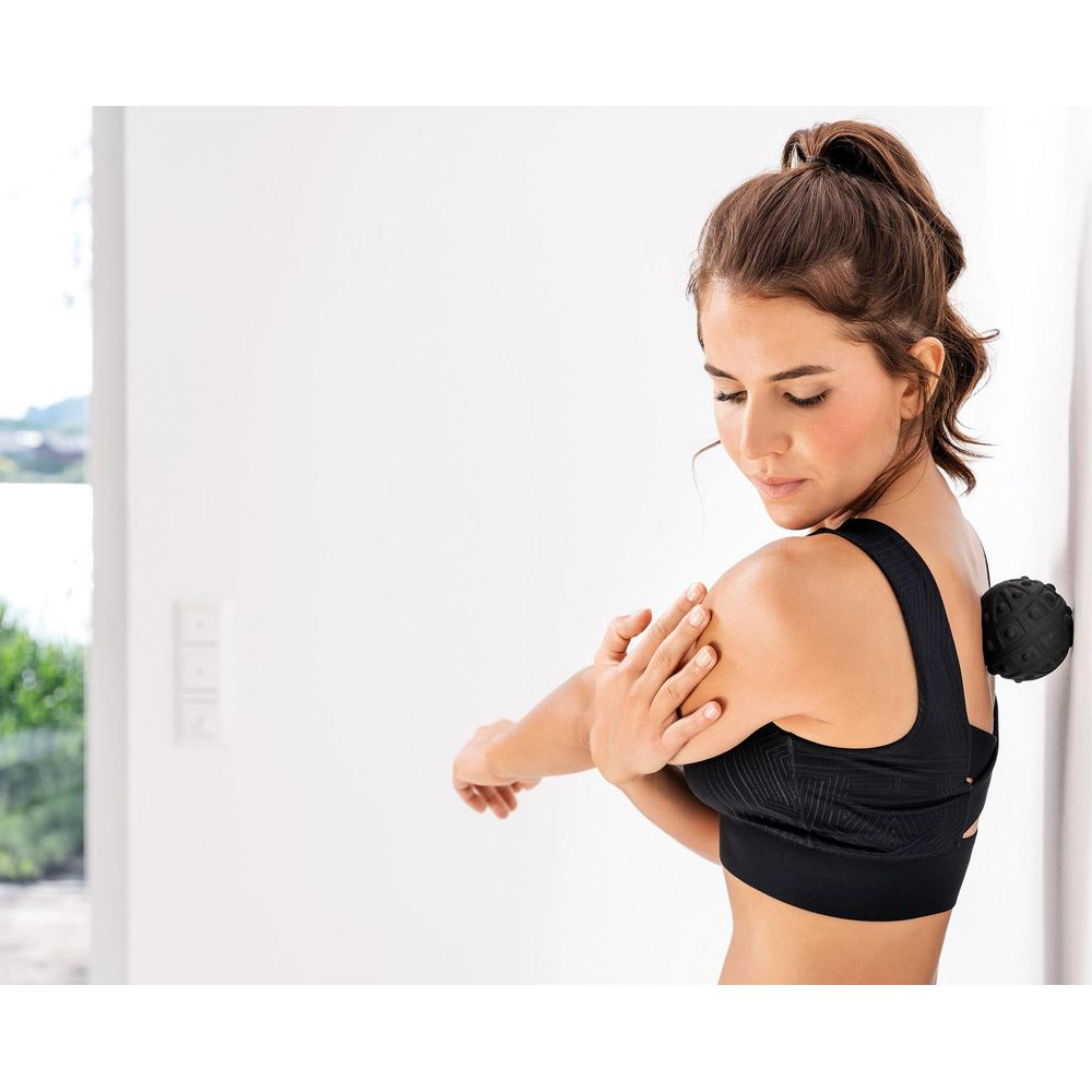 Mujer masajeando su espalda con una pelota negra junto a una pared blanca.