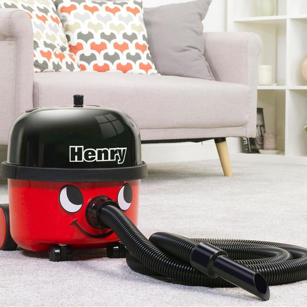 Ein roter Henry-Staubsauger mit schwarzem Schlauch steht auf einem Teppich vor einem Sofa.