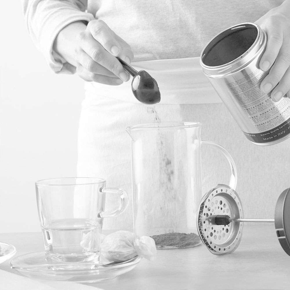 Eine Person schüttet Kaffeepulver aus einer Dose mit einem Löffel in eine Glaskanne.