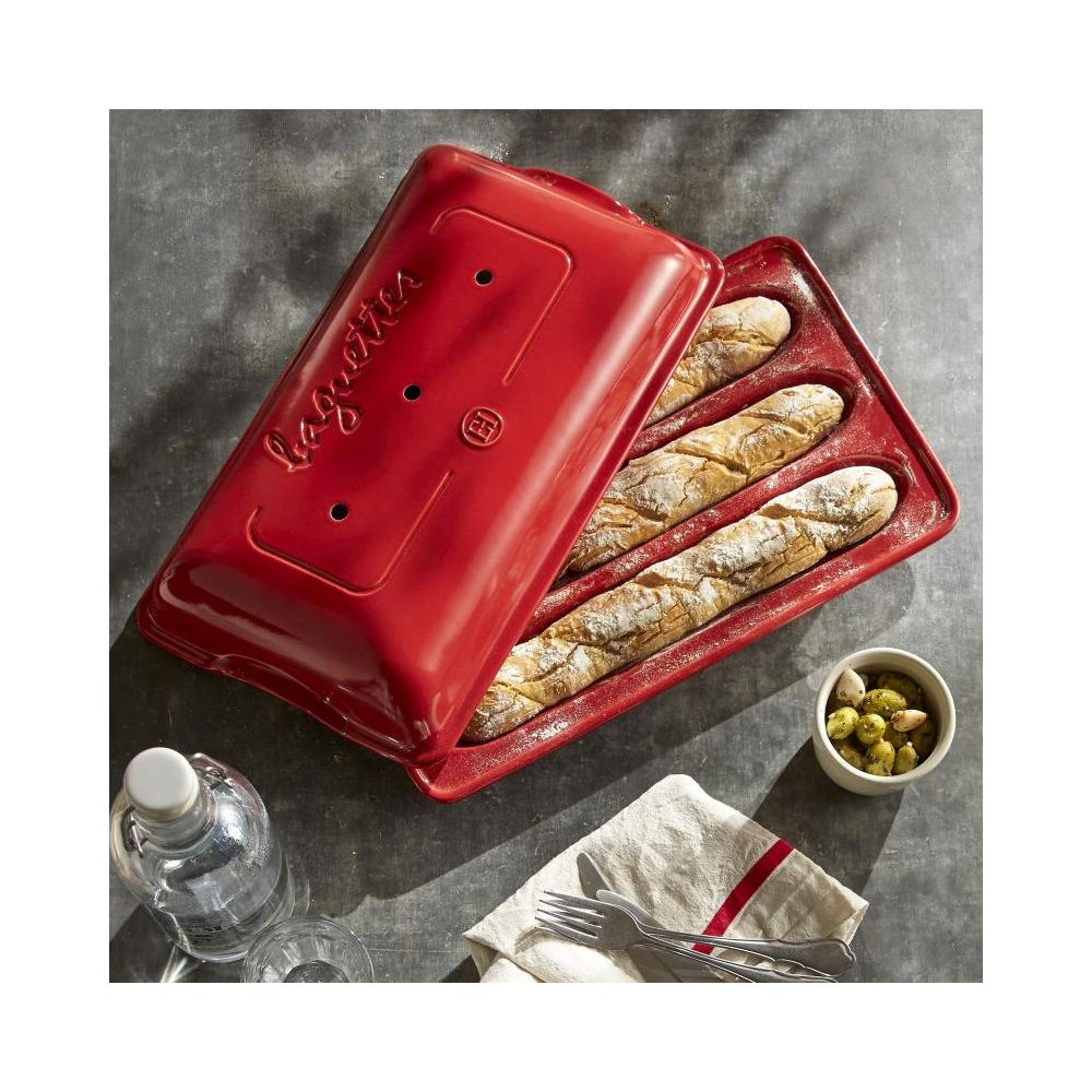 Czerwona forma do bagietek z trzema upieczonymi bagietkami, obok oliwki, woda i sztućce.