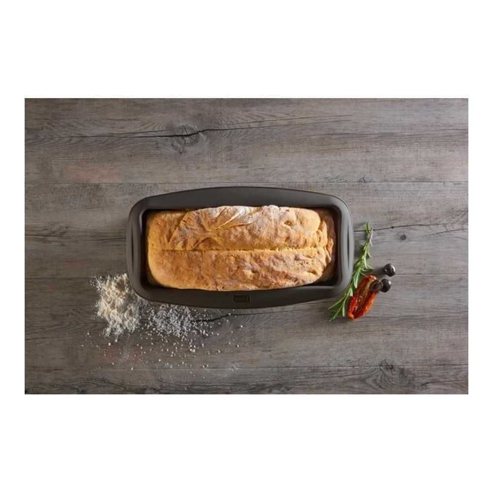 Bochenek chleba w ciemnej formie do pieczenia na drewnianym tle z mąką i dodatkami.