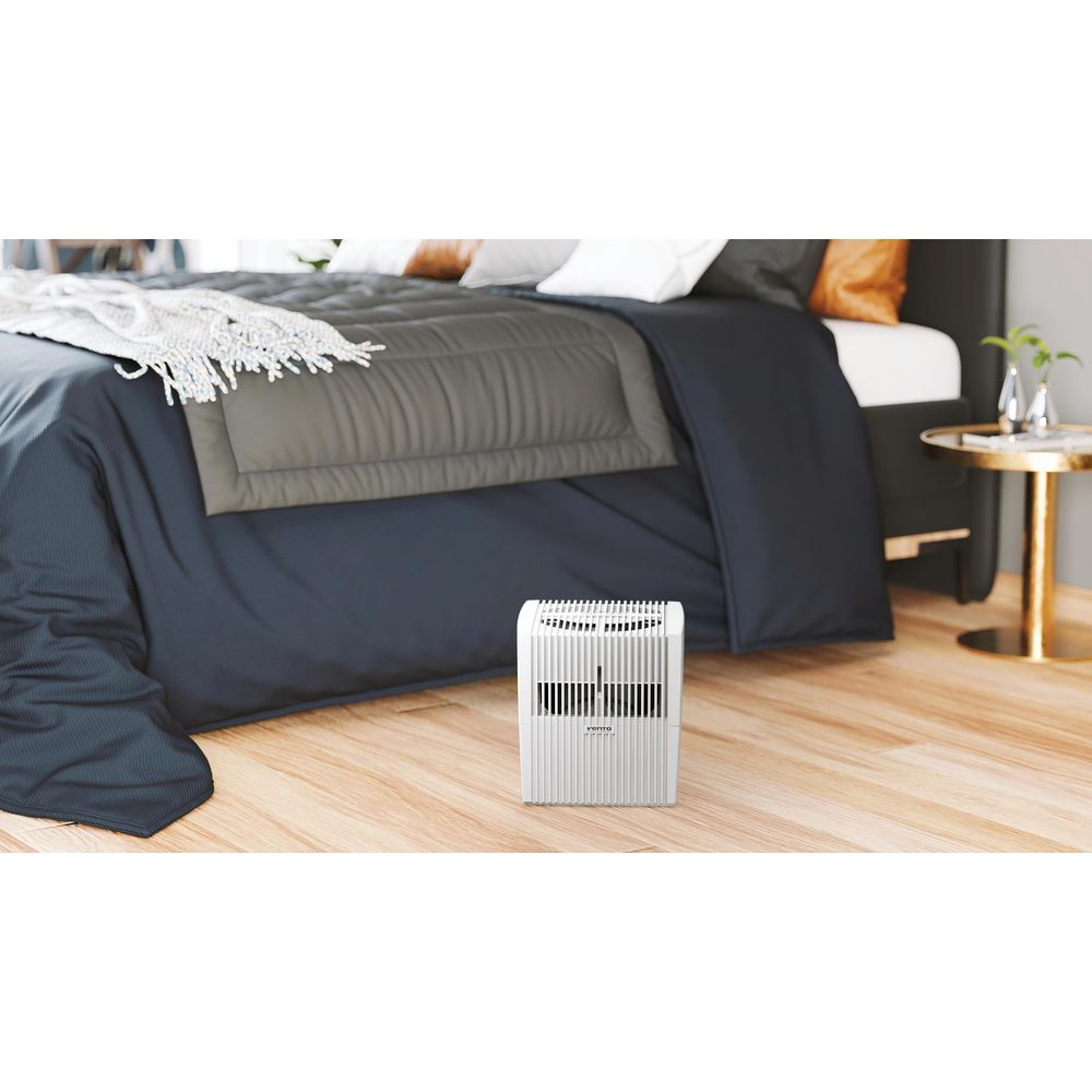 Ein weißer Luftreiniger steht auf einem Holzboden neben einem dunklen Bett.