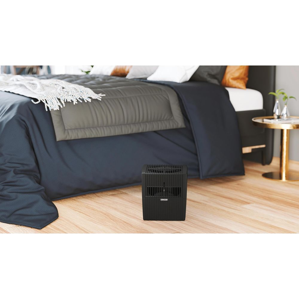 Ein schwarzer Luftreiniger steht auf einem Holzboden neben einem Bett mit dunkler Bettwäsche.