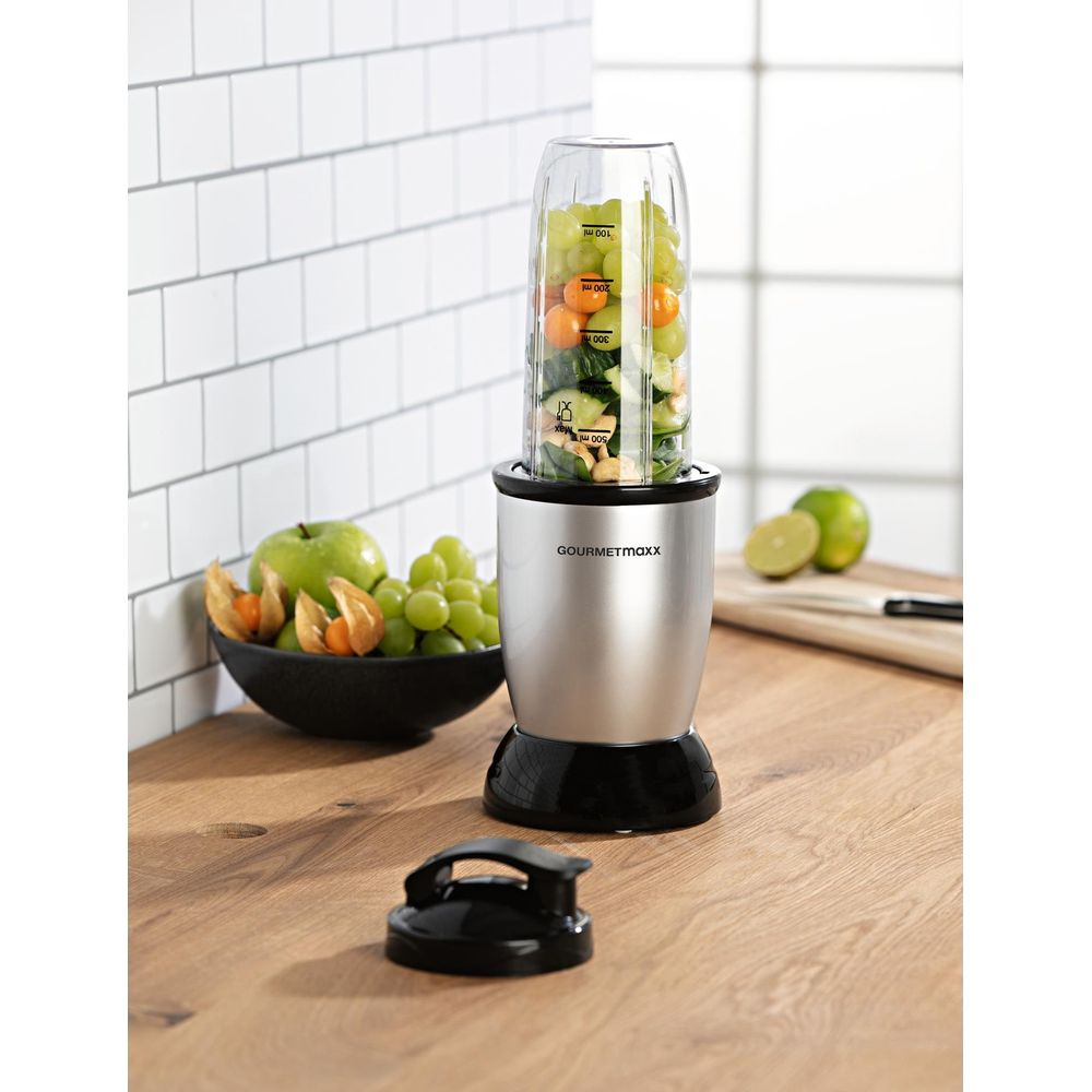 Ein Mixer mit Obst und Grünzeug steht auf einer Theke neben einer Obstschale.