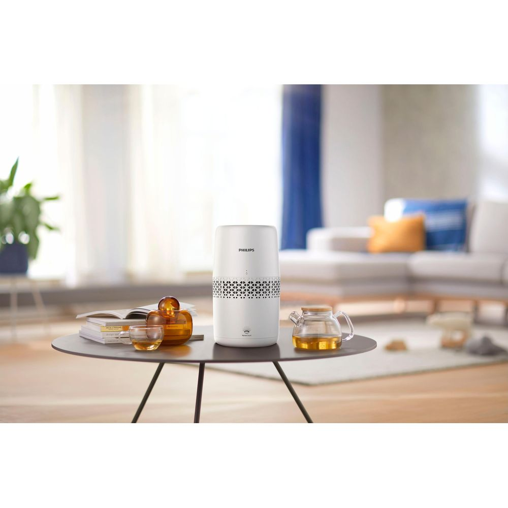 Un purificador de aire Philips blanco está sobre una mesa con libros y vasos de té.