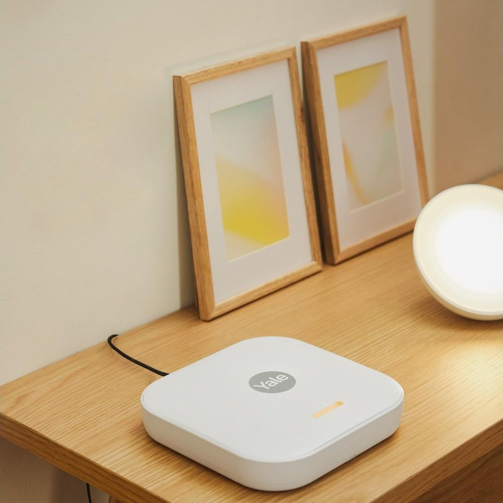 Un dispositivo Yale bianco è su un tavolo di legno con due cornici e una lampada.