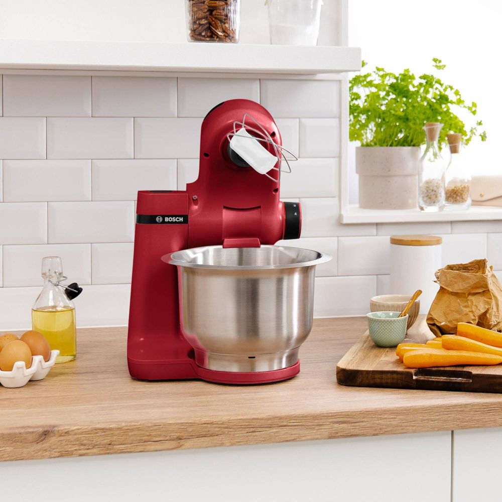 Un robot pâtissier rouge avec bol en inox sur plan de travail en bois près des ingrédients.